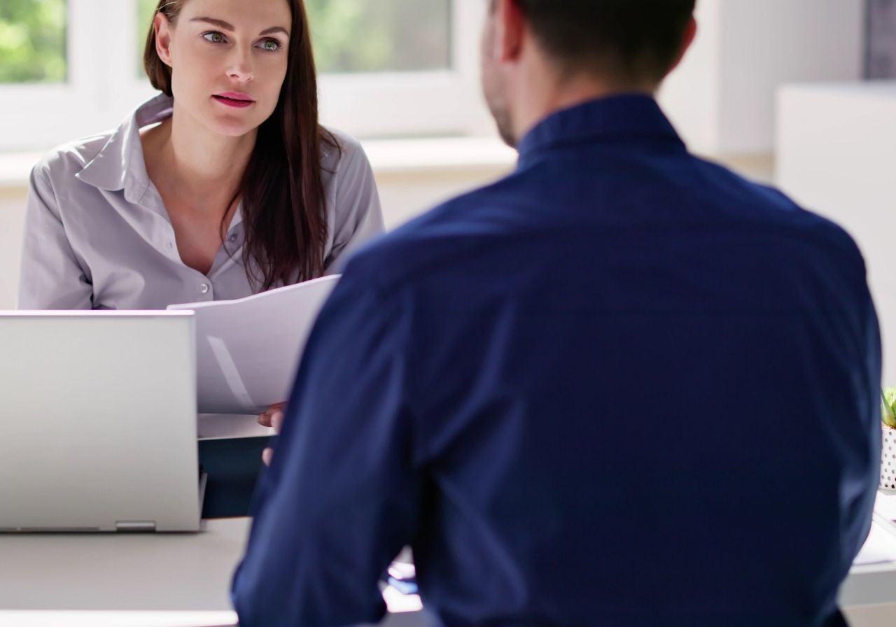 A woman in a light gray blouse is seated at a table, engaging in conversation with a man in a blue shirt, who is facing away from the camera. The woman holds documents and appears to be conducting an interview in a bright, modern office setting.