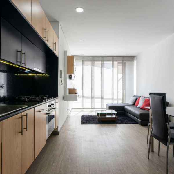 Modern kitchen and living area in a bright apartment, featuring wooden cabinets, a countertop with a stove, a black leather sofa with red cushions, and large windows with sheer curtains.