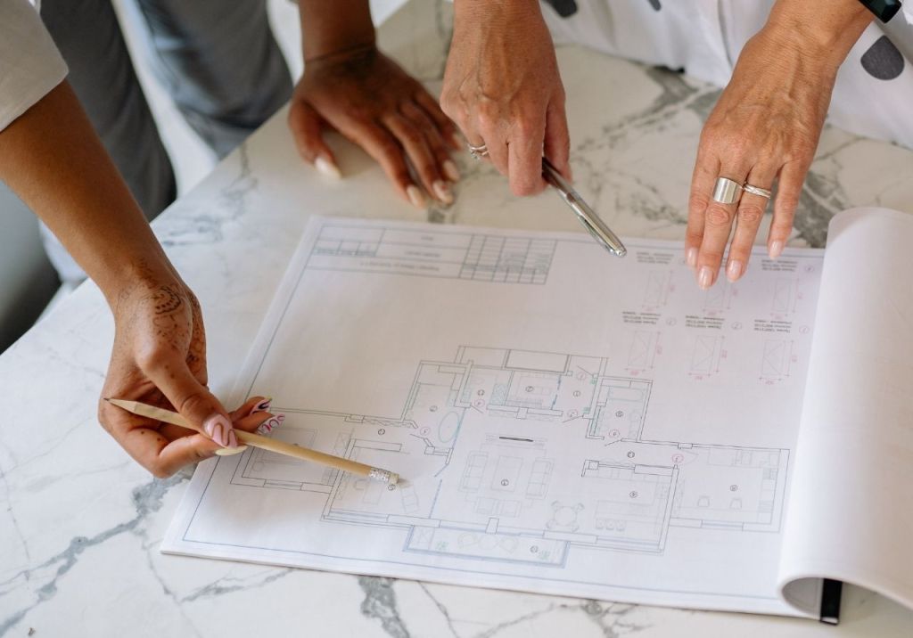Two hands pointing at a floor plan on a marble table, with one hand holding a pencil and the other a pen.