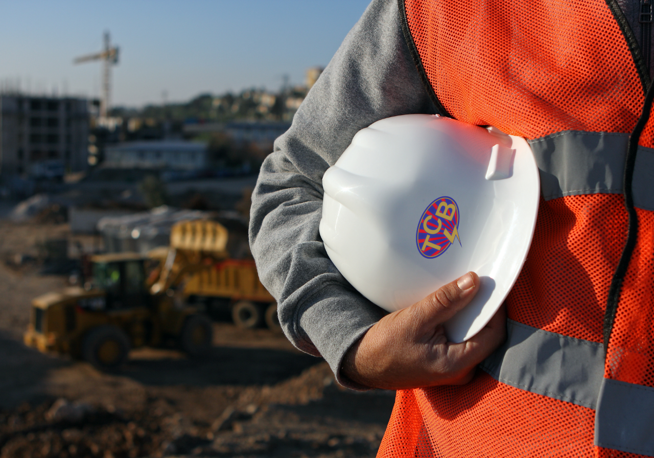 A construction worker in a gray sweatshirt and orange safety vest holding a white hard hat with a logo, with a construction site in the background.