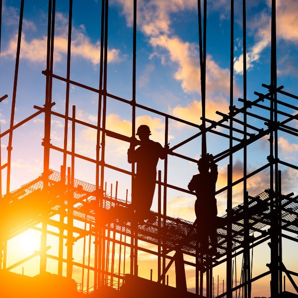 Silhouetted construction workers on scaffolding against a sunset sky with clouds.