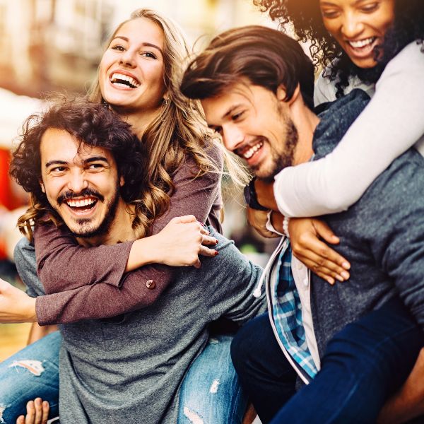A group of four friends joyfully smiling and posing outdoors, with two people giving piggyback rides to two others in a vibrant, sunny setting.