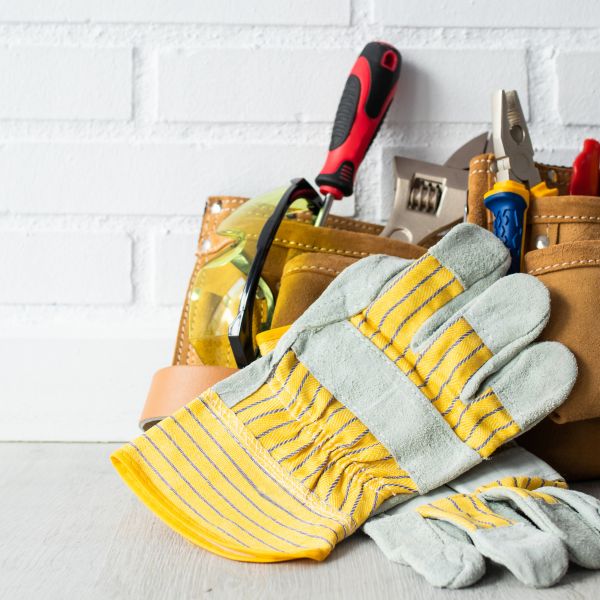 A close-up of a pair of work gloves with yellow stripes, resting on a wooden surface. In the background, a tool belt containing various tools like a screwdriver, pliers, and safety goggles is visible against a white brick wall.