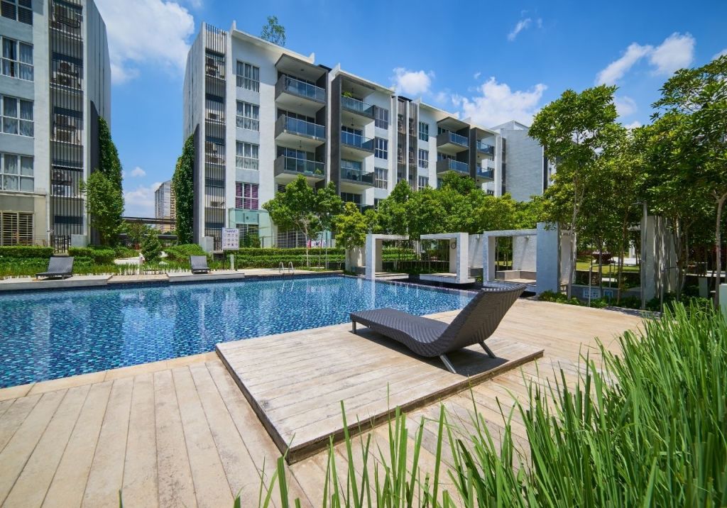 Modern apartment complex with a swimming pool and lounge chairs surrounded by trees and greenery under a sunny sky.