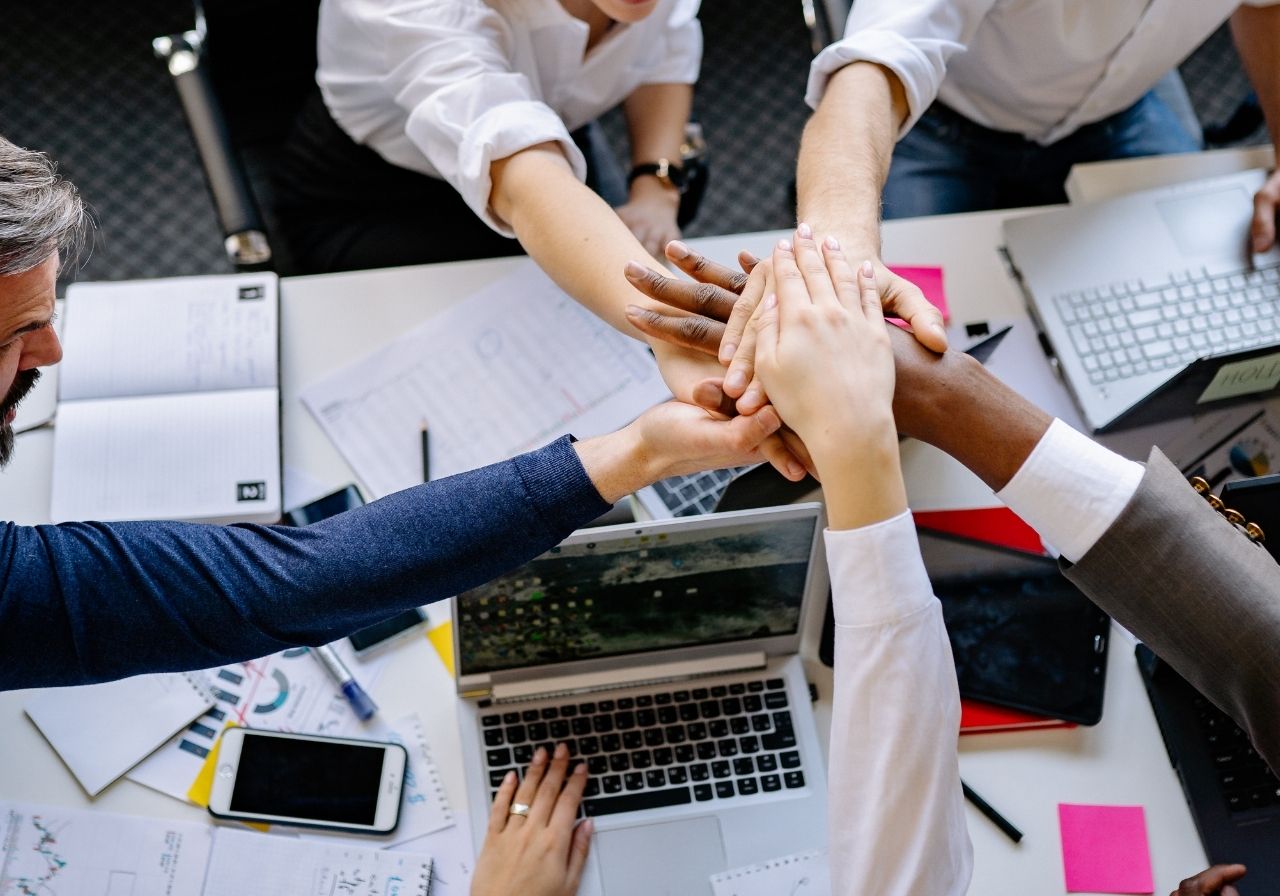 A group of diverse people placing their hands together in a collaborative gesture over a table filled with laptops, notebooks, and paperwork.